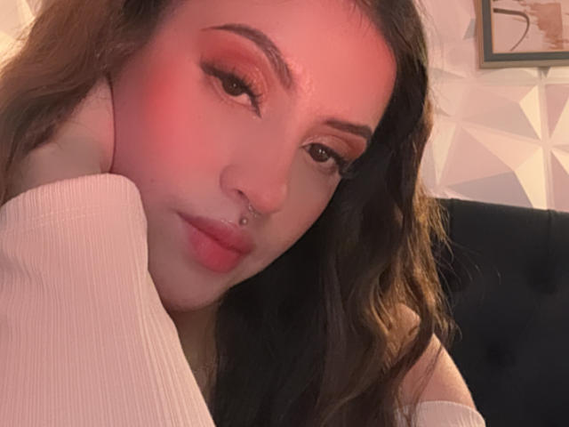 Primer plano de LunaaBlossom con maquillaje de tonos rojos, cejas oscuras y un piercing en la nariz. Está apoyada en su mano, vistiendo una manga blanca, con una expresión sensual. Iluminación suave, fondo desenfocado.