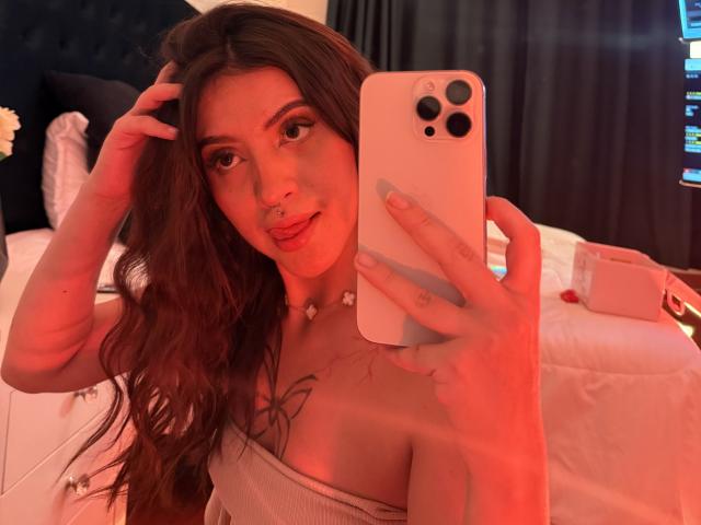 LunaaBlossom with long, wavy brown hair, holding an iPhone, taking a selfie in a red-lit bedroom. She's wearing a strapless white top, hand in hair, seductive gaze, bed in background.