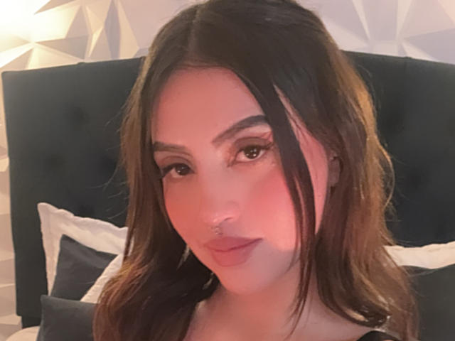 Primer plano de LunaaBlossom con cabello largo y castaño, cejas oscuras y un piercing en la nariz. Ella mira directamente a la cámara con una expresión sutil y seductora. Iluminación suave, cabecera oscura, almohadas blancas.