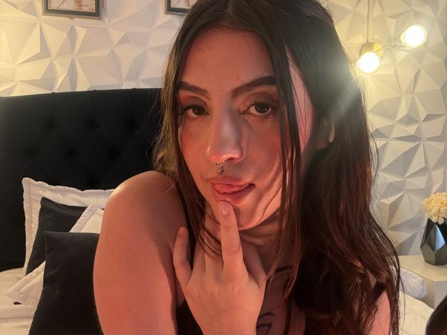 Close-up of LunaaBlossom with long brown hair, nose piercing, and finger on lips. She's wearing a black top, sitting on a bed with white pillows, geometric wallpaper, and a lamp. Fucking sexy.