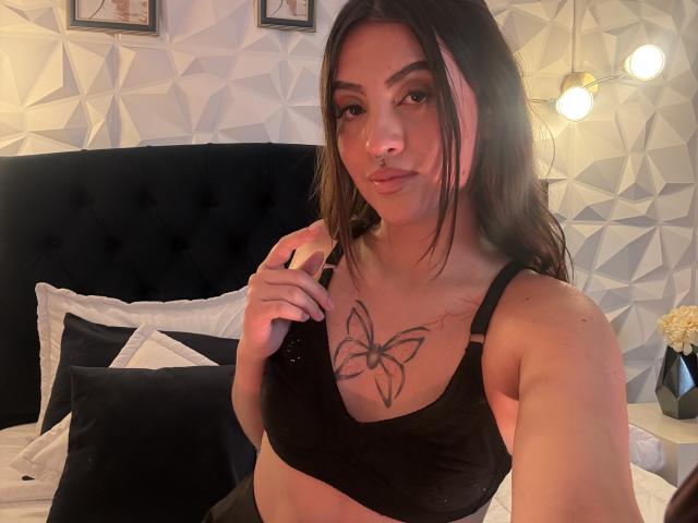 LunaaBlossom, com longos cabelos castanhos, vestindo um sutiã preto, exibindo uma tatuagem de borboleta no peito, sentada em uma cama com almofadas preto e branco, iluminação suave no quarto, olhar sedutor.