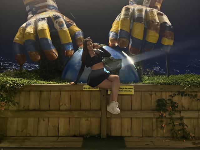 LunaaBlossom sentada en una pared de madera, con las piernas separadas, vistiendo una camiseta corta negra y pantalones cortos, zapatillas blancas, entre dos esculturas gigantes y coloridas con forma de mano por la noche, con las luces de la ciudad al fondo..