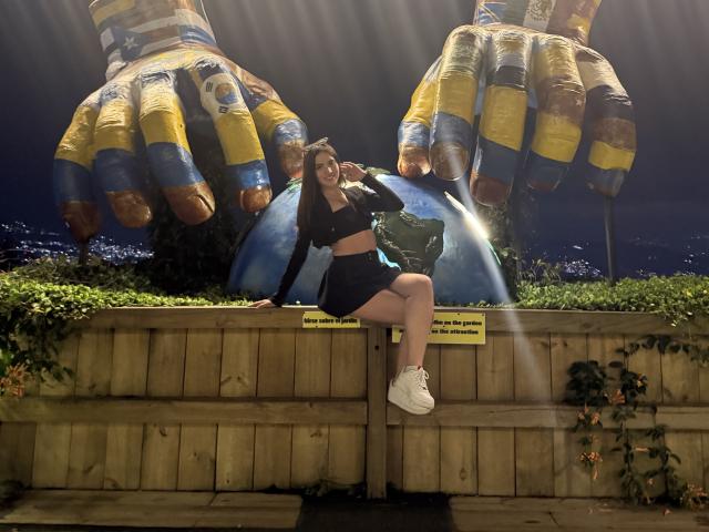 LunaaBlossom, gekleed in een zwart topje en korte broek, zit op een stenen muur met grote, opblaasbare handen in geel en blauw. de achtergrond. Ze heeft witte sneakers aan, en het zonlicht accentueert haar..
