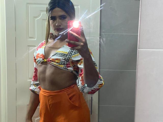 LauraCramz è in piedi davanti a uno specchio del bagno, in un selfie, e tiene in mano un telefono rosso con un flash luminoso. Indossa una canotta colorata che lascia intravedere un addome scolpito e pantaloni arancioni a vita alta..