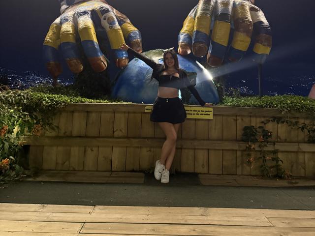 LunaaBlossom leans against a wooden fence at night, wearing a black crop top and shorts, with white sneakers. Large inflatable hands with yellow and blue patterns loom behind her.