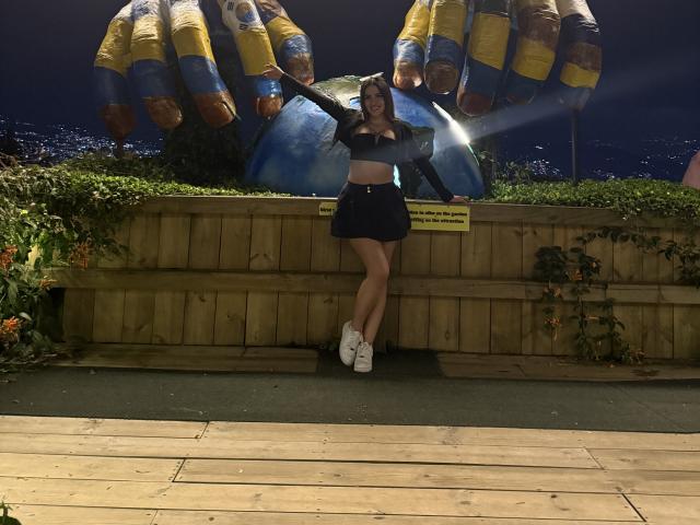 LunaaBlossom stands in front of a wooden wall, leaning against it with legs crossed. She wears a black crop top and mini-skirt, white sneakers. Large blue and yellow inflatable wings behind her.