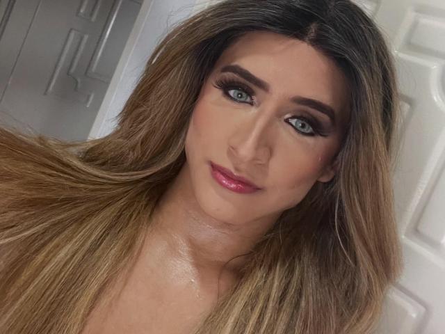 Gros plan de LauraCramz, avec de longs cheveux bruns, des yeux bleus et du rouge à lèvres rose. Elle a des sourcils épais et porte un trait d'eye-liner noir intense. Son expression est légèrement séduisante, avec une pointe de sourire..