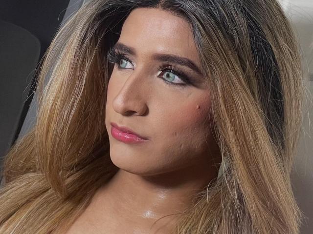 Primo piano di LauraCramz con capelli biondi a metà schiena, occhi azzurri e rossetto rosa. Ha un leggero rossore sulle guance e la pelle luminosa, e distoglie lo sguardo con un'espressione sottile e sensuale..