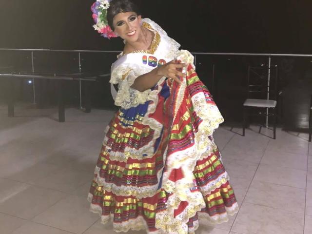 LauraCramz se tient fièrement, vêtue d'une robe traditionnelle colorée et plissée, avec des motifs blancs, rouges et jaunes, et couronne de fleurs.
elle tient la robe, souriant de manière séduisante, avec une tension sexuelle visible..