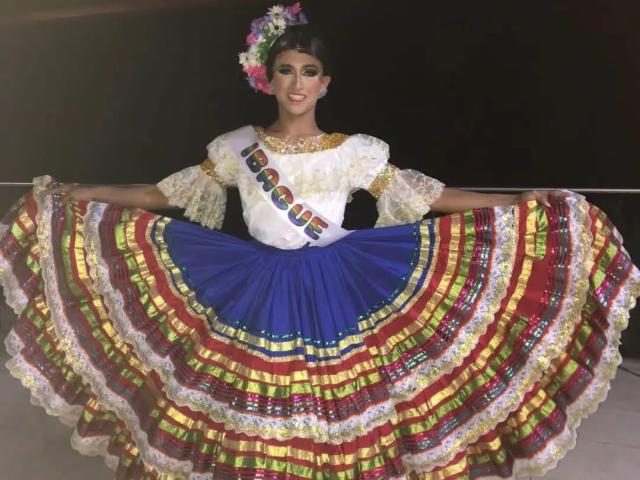 LauraCramz, vêtue d'une robe de flamenco, étale sa jupe colorée et à plusieurs couches. Elle a les cheveux foncés, un serre-tête de fleurs, un chemisier blanc avec des détails dorés, une jupe bleue et un ruban. Tension palpable, sensuelle..