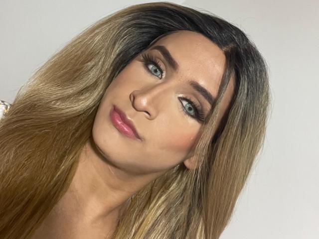 Foto em close-up de LauraCramz, olhando ligeiramente para a direita, com cabelo longo, liso e com efeito ombré, olhos azuis e pele rosada. batom. Sua expressão é confiante, com um toque de sedução. A iluminação intensa realça seus traços..