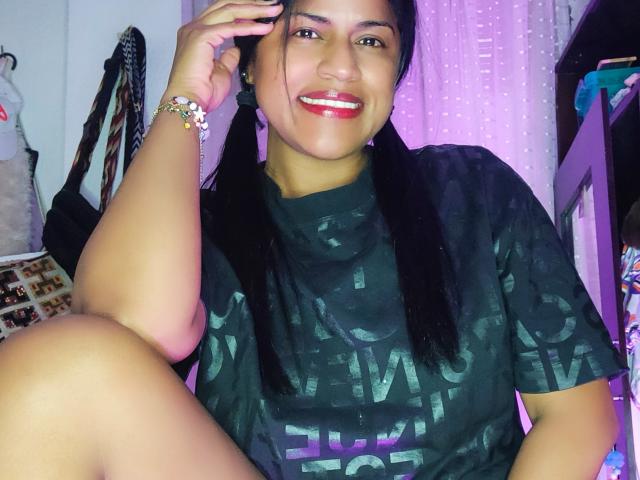 IvannaDuran está sentada com uma perna levantada, sorrindo com batom vermelho. Ela veste uma camiseta preta, usa uma pulseira branca e tem cabelo preto. A mão dela toca a cabeça, criando uma atmosfera sedutora. Luz roxa vibrante..