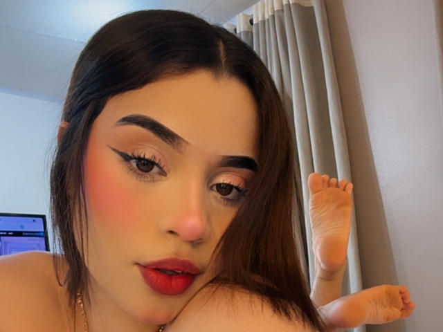 Nahaufnahme von LunaVali, einer attraktiven asiatischen Frau mit langen schwarzen Haaren, rotem Lippenstift und auffälligem Eyeliner, die auf dem Bauch liegt. mit hochgezogenen Füßen, mit einem verführerischen Ausdruck. Fleischlich, erotische Atmosphäre.