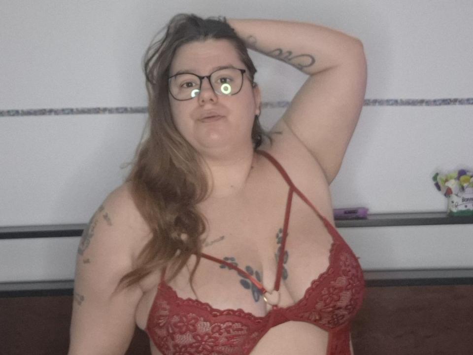 Fotografie von Babyflowers mit heller Haut, braunen Haaren und Brille, die ein rotes Spitzen-BH trägt, der viel Dekolleté zeigt. und Tätowierungen. Ihre rechte Hand ruht auf ihrem Kopf und vermittelt eine sexuelle Spannung..