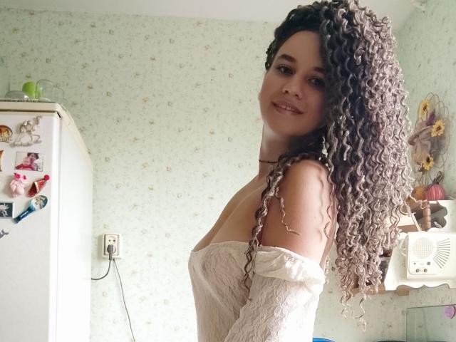 Photographie de SelenaXCute, une femme voluptueuse avec de longs cheveux bruns bouclés, portant un haut blanc en dentelle à épaules dénudées, debout dans une cuisine très lumineuse, elle laissait deviner subtilement ses formes, souriant de manière séduisante..