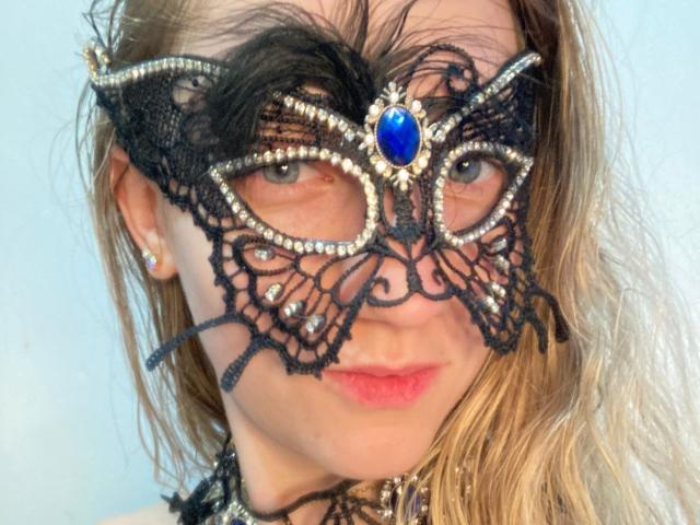 Close-up photo of VenusDivine with a black lace mask adorned with silver beads and a blue gem, covering half her face. She has red lips, light skin, and blonde hair. Fucking sexy.