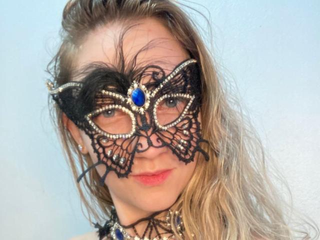 Fotografia ravvicinata di VenusDivine con i capelli biondi spettinati, che indossa una maschera di pizzo nero decorata con dettagli in pietra blu e oro. occhi parzialmente coperti, leggero sorriso, luce naturale soffusa. Incredibilmente attraente..