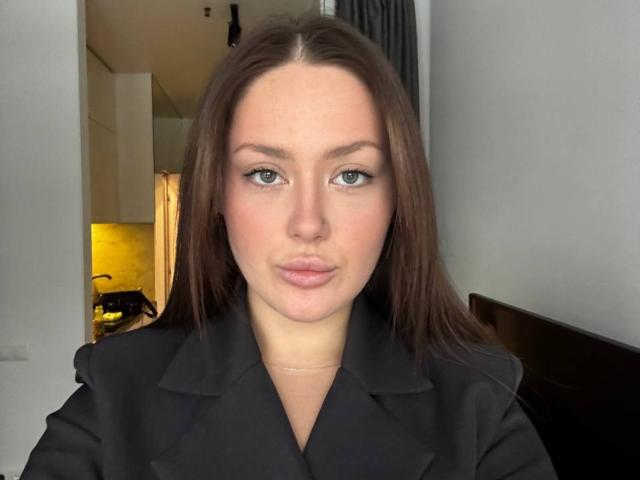 Gros plan d'Elwanda Kiehne, une femme blanche aux longs cheveux bruns, portant un manteau noir, avec une expression légèrement boudeuse et des lèvres d'un rose clair. rouge à lèvres. Expression neutre, maquillage minimal, regard direct, éclairage doux..