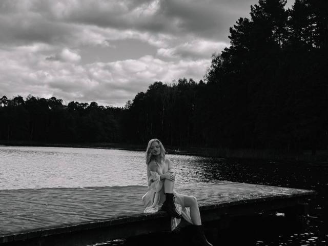 Black and white photograph of CrystalHope, a blonde woman with long hair, sitting on a dark, rocky shore beside a lake. She's gazing at the water, her legs slightly apart.