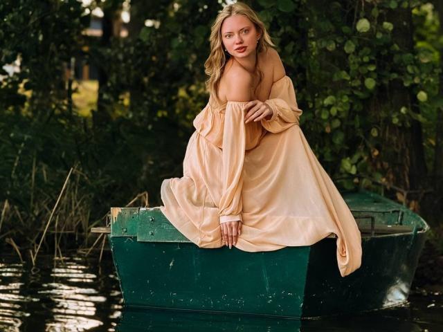 CrystalHope, a blonde woman with fair skin, sits on a green boat in a forest, wearing an off-shoulder, yellow dress that reveals her left breast. Sexual tension, seductive gaze.