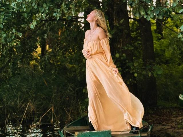 CrystalHope stands on a dock, wearing a flowing, off-shoulder, peach dress, arms behind her back, facing left with eyes closed, sunlight highlighting her blonde hair, forest background. Fucking sexy.