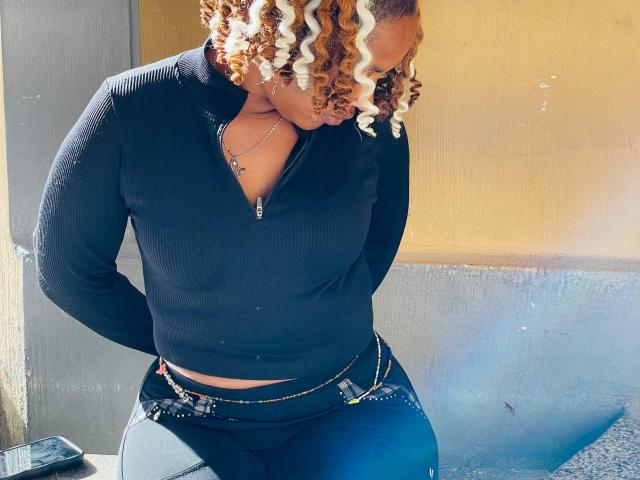 QueenValentina está em pé, com as mãos atrás das costas e a cabeça inclinada, vestindo uma blusa preta com zíper e textura em tiras e calças jeans justas. cabelos loiros e cacheados, a luz do sol realçando suas curvas, fundo de parede azul-acinzentado..