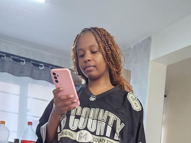 GladTracy, una ragazza di colore con capelli ricci castani, indossa una maglia nera con la scritta "Bounty County", tiene in mano un telefono rosa e guarda in basso. all'opera.
stanza luminosa, tenda grigia, bottiglie d'acqua sullo sfondo..