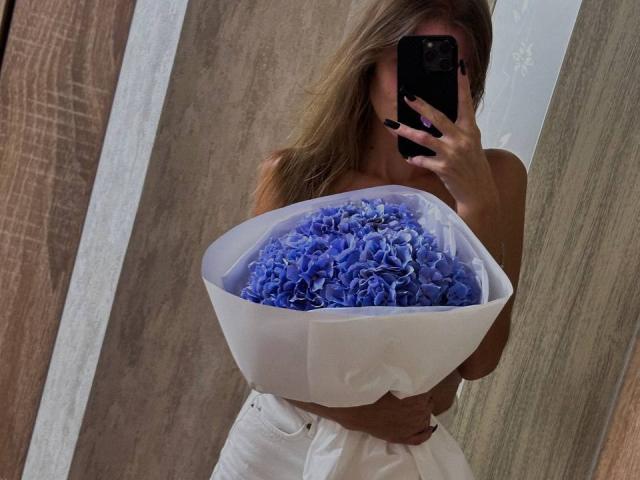 Siyana segurando um grande buquê de flores azuis vibrantes, envoltas em papel branco, tirando uma selfie no espelho com um celular preto. telefone, vestindo um vestido branco. Seu rosto está parcialmente obscurecido..