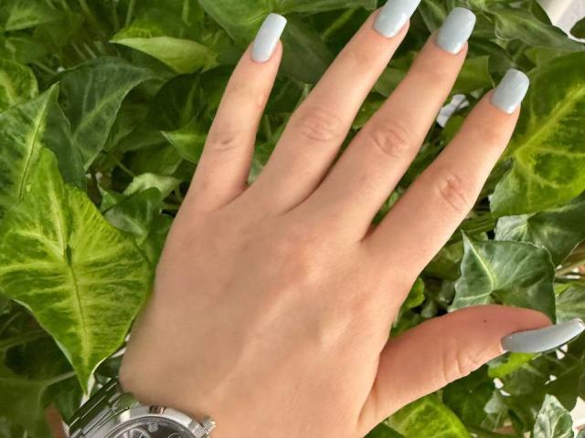 Close-up de uma mão de Siyana, com unhas longas e pontudas em um tom de azul claro, descansando sobre folhas verdes e exuberantes. A pele de Siyana é lisa, e seu pulso está adornado com um relógio de prata. Muito quente.