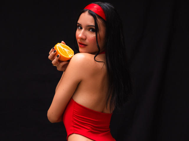 Portrait numérique de LizzeJoy avec des cheveux noirs mouillés, un bandeau rouge et un haut rouge sans bretelles. Elle tient une tranche d'orange près de sa bouche, et regarde par-dessus son épaule avec un regard séduisant..