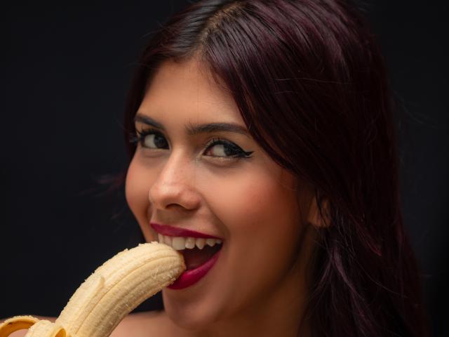 Foto em close-up de DanikaSerov, uma mulher de cabelos escuros com batom vermelho, mordendo uma banana com a boca aberta, mostrando dentes e um leve sorriso. Fundo preto, foco intenso em seu rosto..