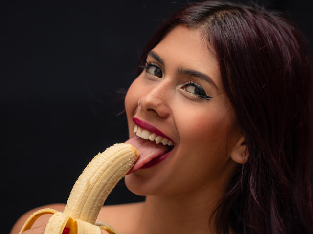 Close-up foto van DanikaSerov, een donkerharige vrouw met een bruine huid, die lacht terwijl ze een gedeeltelijk geschilde banaan aan het aflikken is. Haar tong is uitgestrekt en raakt de banaan aan, waardoor er een erotische spanning ontstaat..