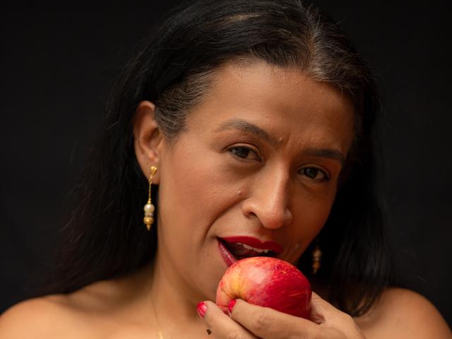 Fotografia em close-up de SoffieMature, com cabelo escuro, segurando uma maçã vermelha perto da boca, mordendo-a levemente. Ela usa brincos de ouro e tem esmalte vermelho. Tensão sexual, expressão sedutora..