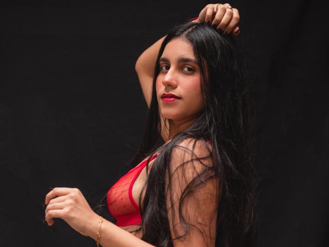 TiffannyRose, eine dunkelhaarige Frau mit langen, nassen Haaren, trägt einen roten Spitzen-BH. Sie hebt eine Hand, klammert sich an ihr Haar und zeigt dabei ihre Brust. Ihr Ausdruck ist verführerisch, dunkler Hintergrund..