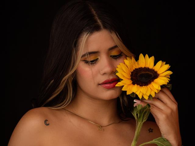 Sheyla Spooner mit langem, dunkelblondem Haar hält einen leuchtend gelben Sonnenblumenkopf dicht vor ihr Gesicht. Sie hat gelben Lidschatten, einen Nasenring und Tätowierungen an ihrem Schulterblatt. Schwarzer Hintergrund. Verdammt sexy..