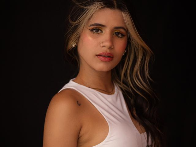Fotografie von Sheyla Spooner, die frontal aufgenommen wurde, mit einem weißen Top ohne Ärmel, langen blonden Haaren, braunen Augen und rosafarbener Haut. Lippen, feine Sommersprossen und einen kleinen Muttermal auf ihrer linken Schulter. Dunkler Hintergrund. Sexuelle Spannung..