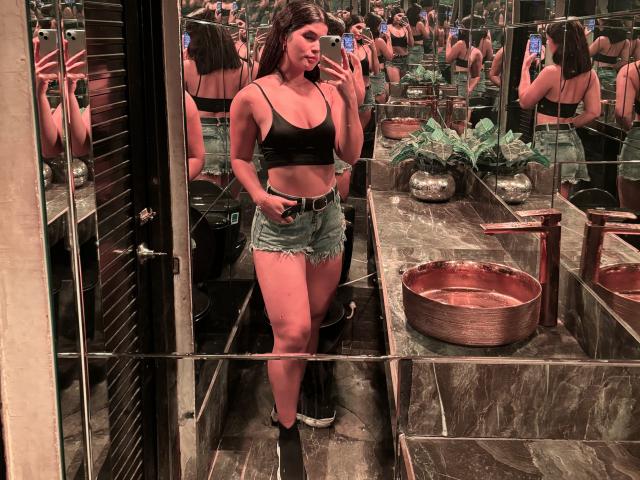 ScarlletZhen, a curvy woman with dark curly hair, wears a black crop top and denim shorts, holding a phone, walking through a mirror-lined, dimly-lit bathroom with a red basin. Fucking sexy.