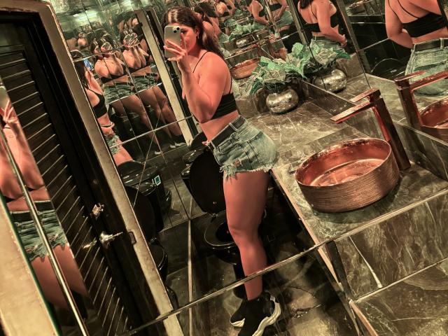 ScarlletZhen, a topless woman with dark hair, stands in a dimly lit, reflective room wearing shiny green shorts, showing a toned body and ample breasts. Multiple reflections highlight the scene.