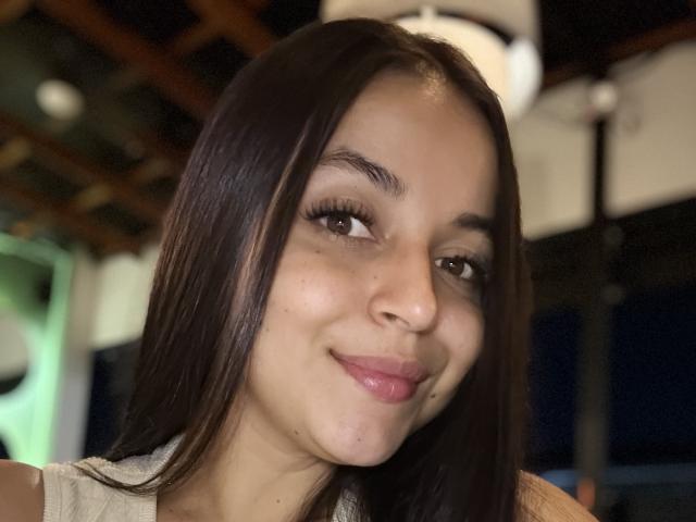 Close-up of HikariChloe with long dark hair, light skin, and full pink lips. She's smiling slightly, looking off to the side. Soft lighting, blurred background. Slight sexual tension.