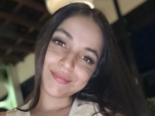 Close-up photo of HikariChloe with dark, wavy hair, light skin, and brown eyes, smiling slightly. Soft light highlights her face, casting shadows. She has a nose ring and subtle makeup.