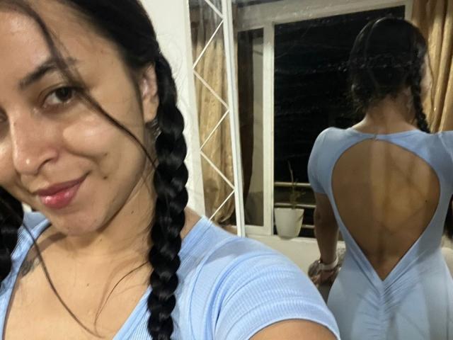 Fotografía de primer plano de HikariChloe, una mujer de piel morena con cabello trenzado, que lleva una blusa azul claro con un escote pronunciado en la espalda y está mirando. mirando a la cámara con una sonrisa sutil. La vista posterior muestra la parte trasera abierta..