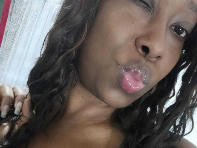 Fotografía de primer plano de EbonyAngelForU con piel oscura, cabello negro ondulado y labios rosados brillantes ligeramente fruncidos en un beso. Ella tiene ojos oscuros, uñas largas y una ligera sonrisa. Increíblemente sexy..
