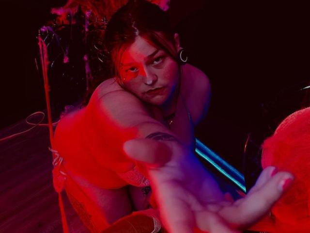 MilanDallila, een bezwete, naakte vrouw met donker haar, hurkt diep, reikt vooruit met een uitgestoken vinger, badend in rood en blauw neonlicht.
intense, seksuele spanning, donkere achtergrond.