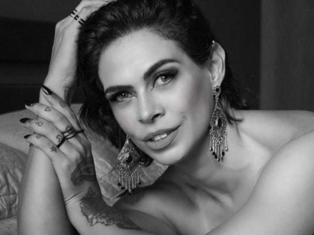 Photographie en noir et blanc de VeronnaInk, allongée sur un lit, la tête penchée, une main derrière la tête, portant de grandes boucles d'oreilles pendantes. boucles d'oreilles, poitrine nue, tatouage sur le bras gauche, sourire narquois, teint bleuté, séduisant, regard intense.