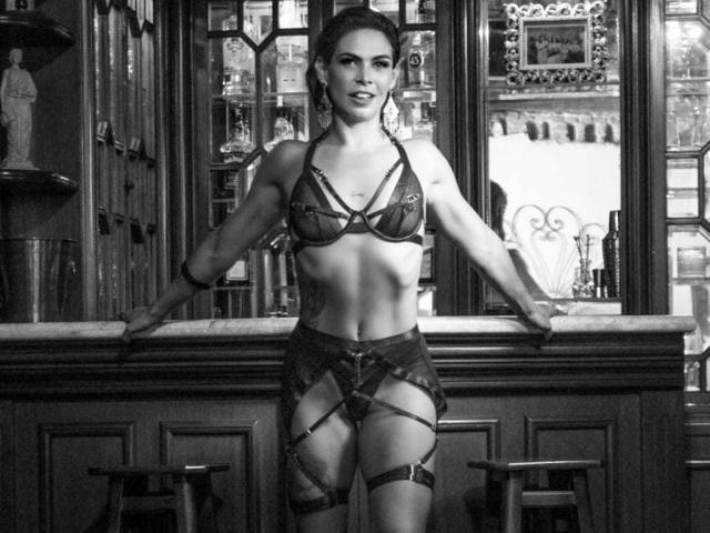 Photo en noir et blanc de VeronnaInk, debout dans un bar, portant de la lingerie à fines bretelles qui expose sa petite poitrine. Corps tonifié.
elle se penche contre le bar, les mains sur le comptoir, avec un regard séduisant..