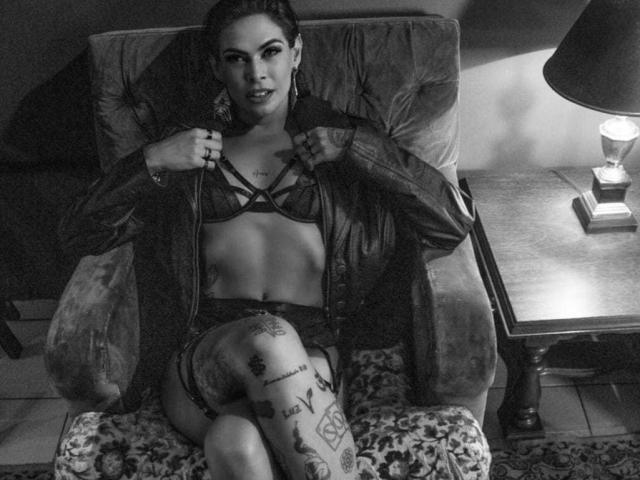 Foto em preto e branco de VeronnaInk, sentada em uma cadeira estampada, vestindo lingerie com alças finas e um casaco aberto. exibindo tatuagens em sua coxa. Ela está olhando diretamente para a câmera com uma expressão sedutora. Lâmpada à direita.