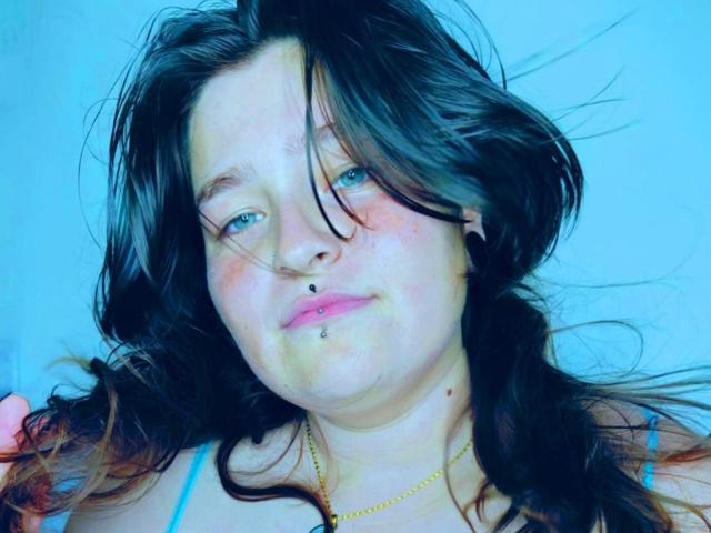 Close-up of MilaDallila with long, messy black hair, blue eyes, and a piercing on her lower lip. She has a pale complexion and a subtle smile. Bright blue background. Fucking sexy.