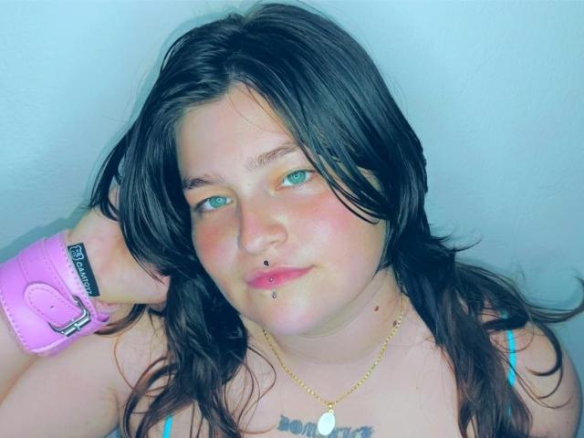 Close-up van MilaDallila met rommelig zwart haar, blauwe ogen en een lip-piercing. Ze draagt een roze polsbandje en een gouden ketting met de tekst "maan". Ze kijkt recht in de camera met een enigszins verleidelijke uitdrukking..