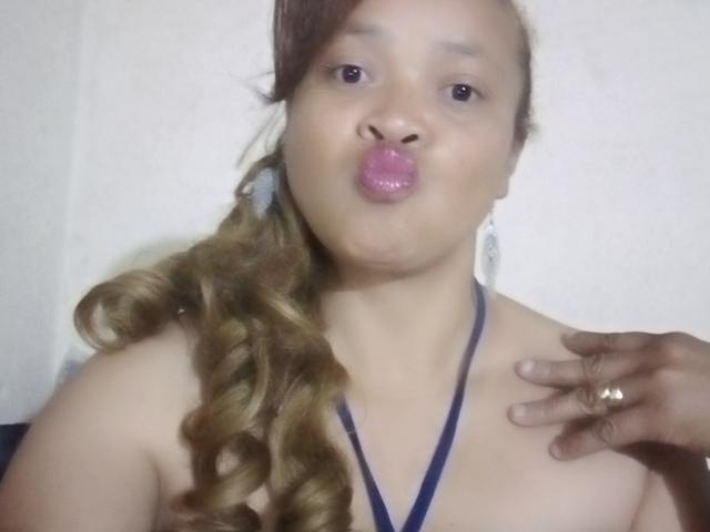 Fotografía de Rahel69, una mujer asiática con sobrepeso, de piel marrón claro y cabello rizado, que frunce sus labios rosados y brillantes, y que lleva puesta una camiseta negra con cuello halter, con la mano derecha tocando su clavícula..