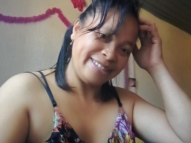 Photographie de Rahel69, souriante, avec les cheveux noirs mouillés, touchant sa tête avec une main. Elle porte un haut à bretelles fines avec un motif floral. L'arrière-plan comprend des guirlandes roses et une fleur rouge..